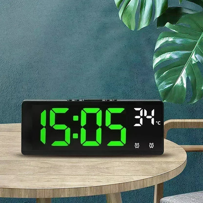Digital clock with green display on a wooden table against a teal wall with a plant.