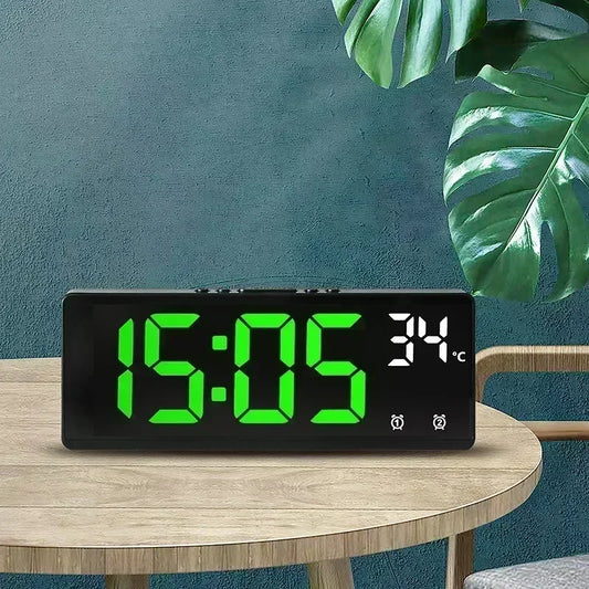 Digital clock with green display on a wooden table against a teal wall with a plant.